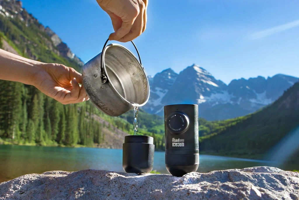 ریختن آب از قابلمه فلزی داخل مخزن یک اسپرسوساز دستی قابل‌حمل کنار فنجان کوچک، روی صخره با پس‌زمینه دریاچه و کوه‌های برفی.