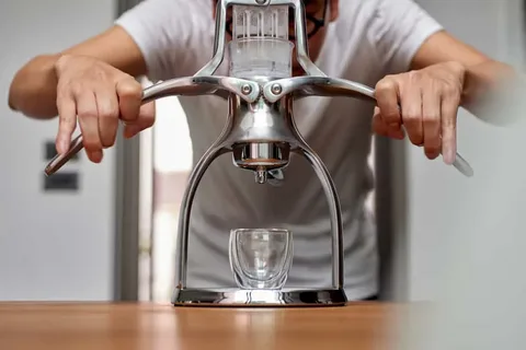 نمای نزدیک از یک نفر که با دستگاه اسپرسوساز دستی اهرمی در حال گرفتن قهوه روی یک فنجان شیشه‌ای کوچک روی میز چوبی است.