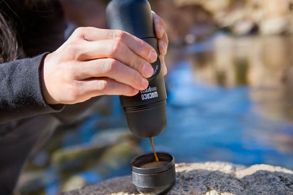 ریختن قهوه از اسپرسوساز دستی قابل‌حمل مشکی داخل فنجان کوچک در فضای باز کنار رودخانه.
