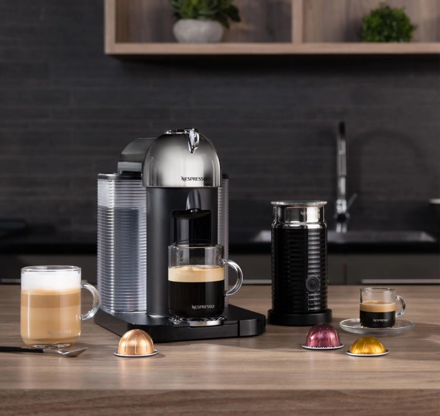 دستگاه قهوه‌ساز کپسولی نسپرسو روی کانتر آشپزخانه به‌همراه فوم‌ساز شیر، فنجان‌های قهوه و چند کپسول رنگی در جلوی دستگاه.