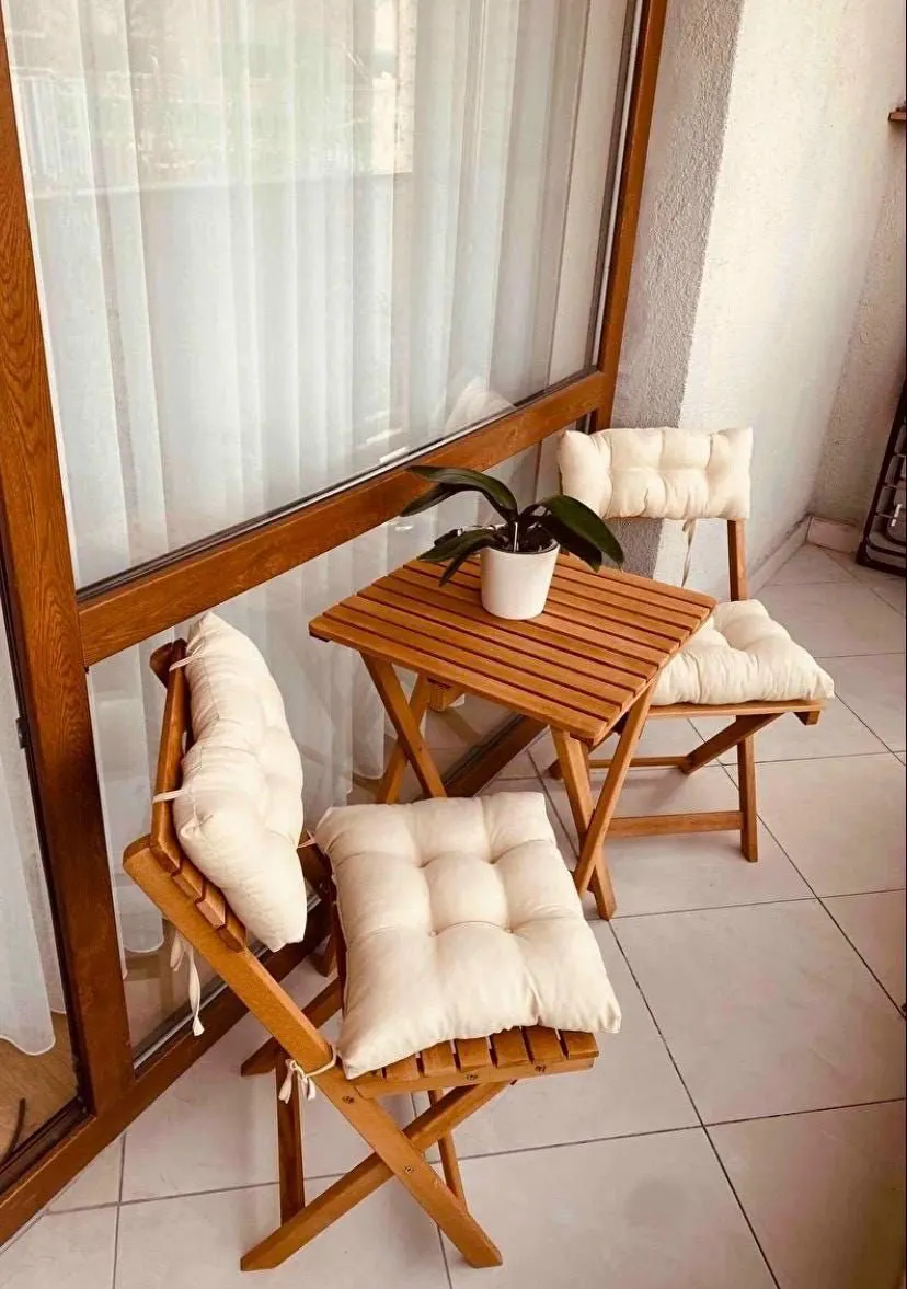 بالکن کوچک با میز و دو صندلی تاشوی چوبی و کوسن‌های سفید، همراه گلدان گیاه روی میز کنار پنجره بزرگ.